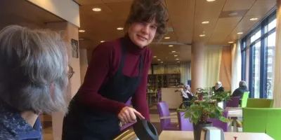 Vrouw zit aan tafel en krijgt koffie ingeschonken door medewerker
