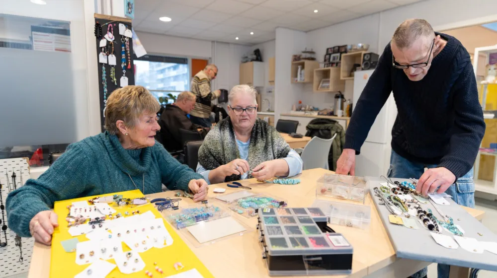 Twee vrouwen zitten aan tafel en maken sieraden en man staat er bij