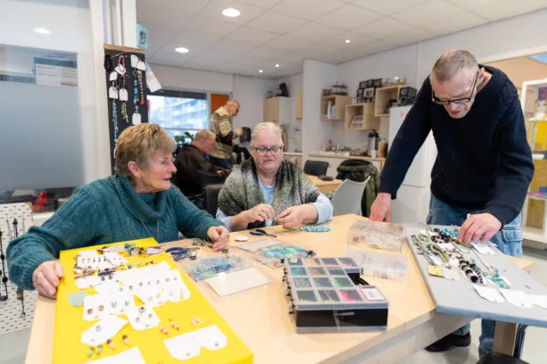 Twee vrouwen zitten aan tafel en maken sieraden en man staat er bij