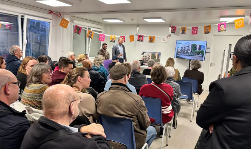 Een groep mensen in een bouwkeet, krijgen een presentatie over een nieuwbouw gebouw. Er hangen vlaggetjes met gezichten er op van de mensen die in de nieuwe appartementen gaan wonen.
