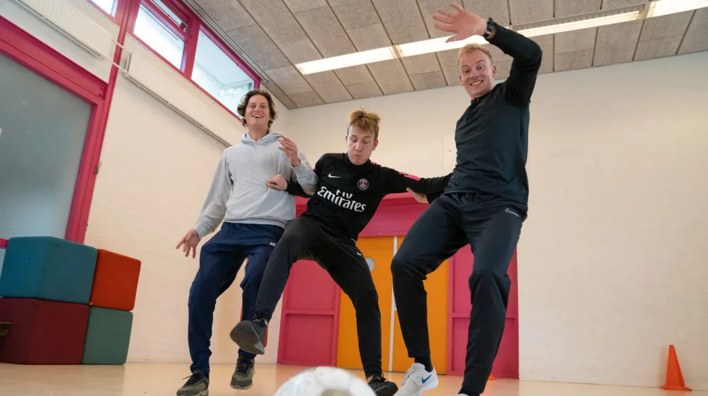 Twee sportcoaches van Pameijer en een jongen voetballen binnen in een sportzaal en hebben plezier