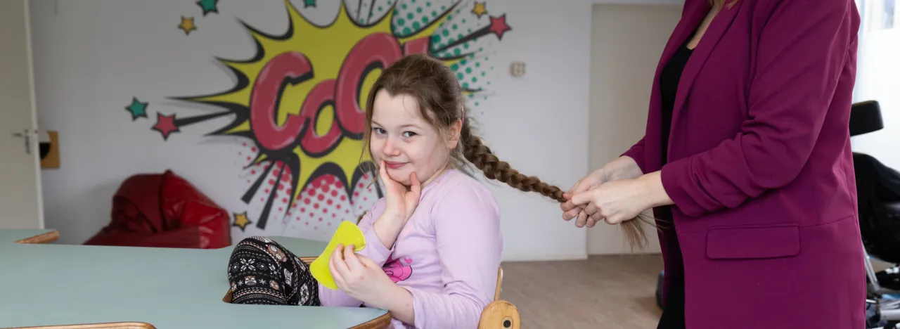 Kind zit aan tafel en kijkt de camera in terwijl iemand haar haren vlecht