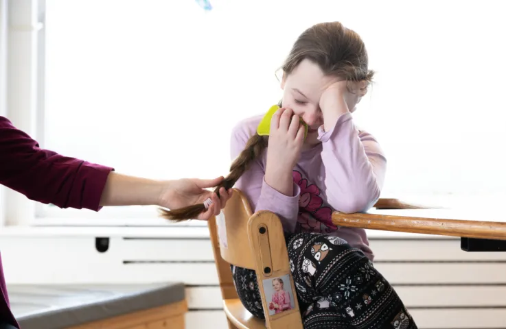 Kind zit aan tafel met een hand voor het gezicht terwijl iemand haar vlecht vasthoudt