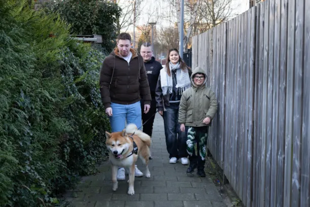 Een groepje van drie kinderen en een wooncoach laten de hond uit, ze lopen langs een schutting en een heg