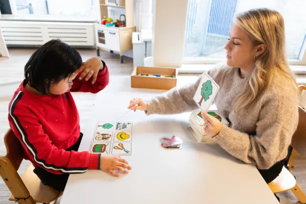 Kind en medewerker zitten aan tafel en spelen een spel met figuren