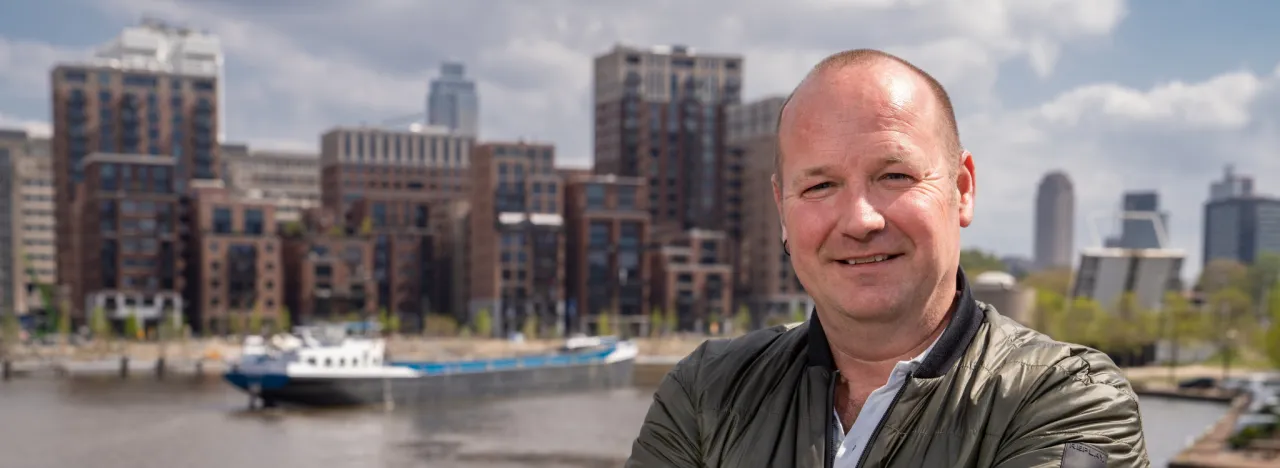 Man poseert met armen over elkaar voor de camera op een brug met op de achtergrond de stad