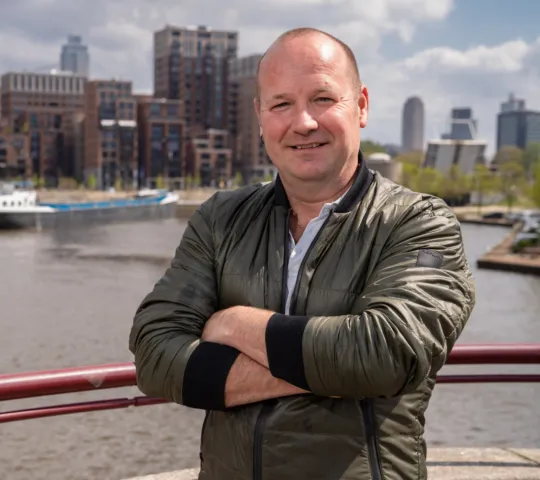 Man poseert met armen over elkaar voor de camera op een brug met op de achtergrond de stad