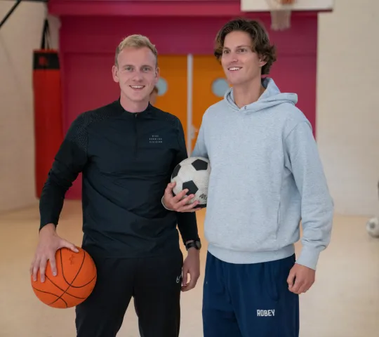 Twee medewerkers poseren met een bal onder de arm in een gymzaal