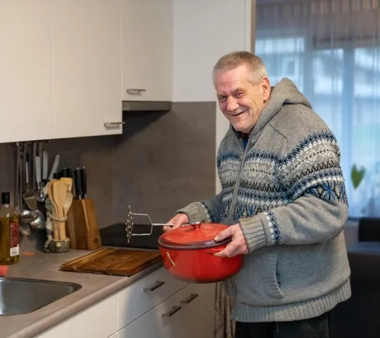 Man staat met een pan in de hand in een keuken en lacht naar de camera