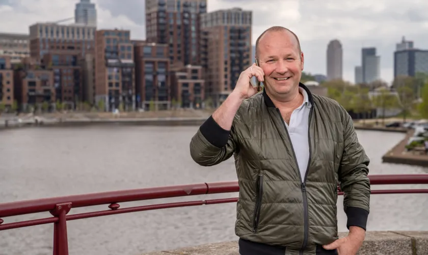 Man staat bellend op een brug en lacht naar de camera met achter zich de stad