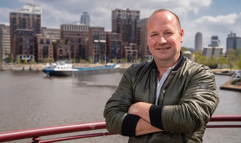 Man poseert met armen over elkaar voor de camera op een brug met op de achtergrond de stad