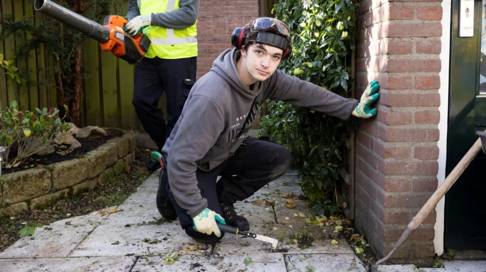 Man zit gehurkt en is onkruid aan het wieden terwijl hij de camera inkijkt