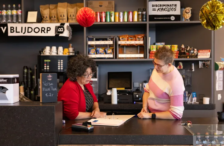 Twee vrouwen staan achter de bar van voetbalclub Blijdorp. De ene vrouw kijkt in een map en legt iets uit aan de andere vrouw