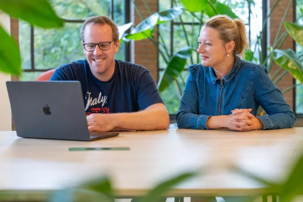 Twee personen zitten aan tafel en kijken samen lachend naar de laptop