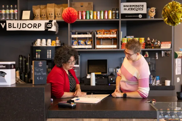 Twee vrouwen staan achter de bar van voetbalclub Blijdorp. De ene vrouw kijkt in een map en legt iets uit aan de andere vrouw