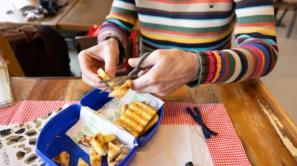 Iemand met een gestreepte trui knipt brood in stukjes in een blauwe broodtrommel