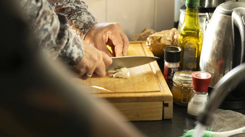 Iemand snijdt met een groot mes een bol knoflook, eromheen staan kruiden en olijfolie