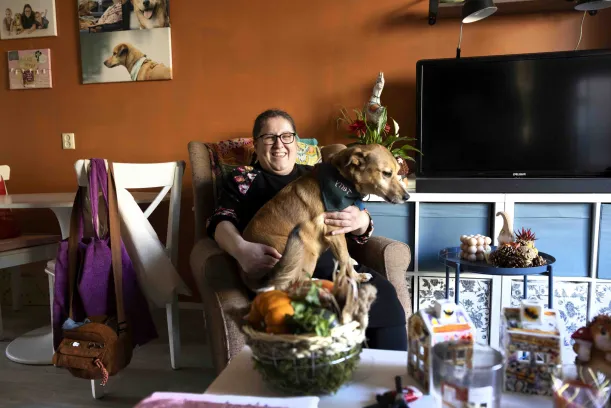 Een vrouw zit in haar huiskamer, ze heeft haar hond op schoot en kijkt blij